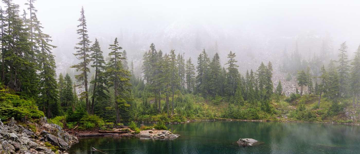 Mist over Melakwa Lake in the Alpine Lakes Wilderness — dark conifers and pale scree rising into fog above still green water.